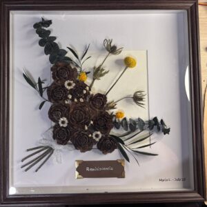 Cuadro botánico en marco de profundidad con piñas de pino en forma de rosa, eucalipto, cardos secos, craspedia y paniculata sobre fondo crema. Pieza firmada a mano por María E., julio 2026.